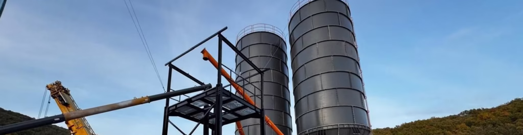Twin bolted kitset silos with modular steel construction installed at an industrial site with crane support, optimized for bulk cement and mineral storage.