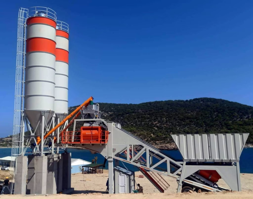 Mobile concrete batching plant with dual vertical cement silos, conveyor system, and aggregate hopper on a coastal construction site.